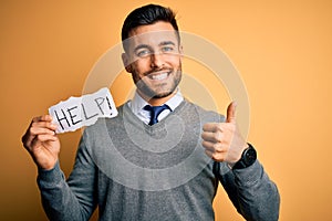 Young overworked business man asking for help holding paper over yellow background happy with big smile doing ok sign, thumb up