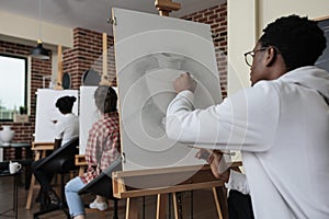 Young painter sitting in front of white canvas drawing vase model