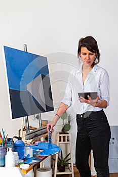 The young painter is standing in her home studio and copying her work from her tablet directly on the canvas
