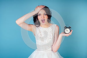 Young and panic beautiful woman bride with makeup in white wedding dress holds an alarm clock in hand and screams