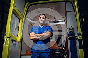 Young paramedic in a blue uniform standing and smiling in front of an an ambulance car
