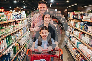 Young parents and daughter in grocery store. They walk among hygeine shelfs and smile on camera. Girl carry trolley. Man