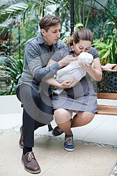 Young parents holding their newborn infant, kissing and looking at baby, sitting on bench indoor