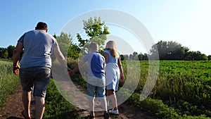 Young parents with little son hold hands of each other walking along rural road near green meadows. Mother and father