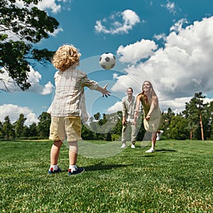 Young parents playing footabll with their little son on grass field in the park on a summer day