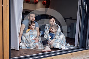 Young parents and small children sit on floor of living room at home in front of open panoramic window