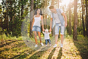 Young parents are taught to walk a daughter of one year on nature in a park on a lawn. The first steps of the child`s hand with