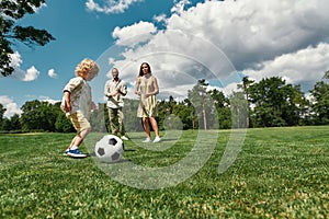 Young parents teaching their little son kicking football on grass field in the park on a summer day