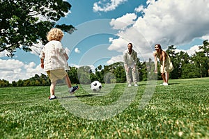 Young parents teaching their little son playing football on grass field in the park on a summer day
