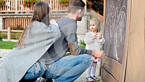 Young parents with their cute little girl drawing on blackboard. Child holding chalk and drawing.