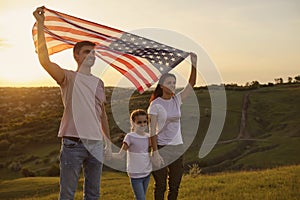 Young parents with their daughter holding American flag in countryside at sunset. Independence Day celebration