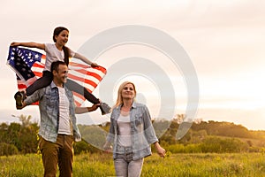 Young parents with their daughter holding American flag in countryside at sunset. Independence Day celebration