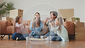 Young parents and two daughter enjoying hot fresh pizza, sitting together on floor among cardboard boxes after moving