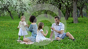 Young parents with two daughters spend leisure on a plaid in park on spring