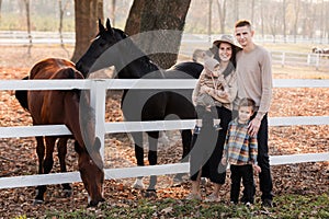 Young parents walk with little daughter and son near horses in autumn park. Dad and mom holding baby girl. Happy family concept