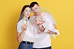 Young parents wearing white casual t shirts posing with new born daughter, mother showing toy for little baby with nippel,