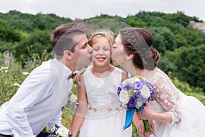 Young parents in wedding dresses kiss their young daughter in cheeks