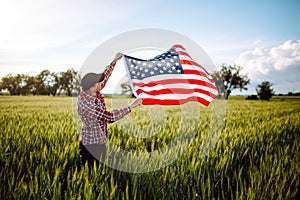 Young patriotic farmer stands among new harvest. Boy walking with the american flag on the green wheat field celebrating national