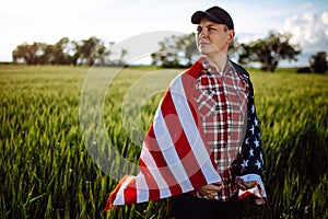 Young patriotic farmer stands among new harvest. Boy walking with the american flag on the green wheat field celebrating national