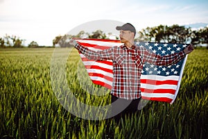 Young patriotic farmer stands among new harvest. Boy walking with the american flag on the green wheat field celebrating national