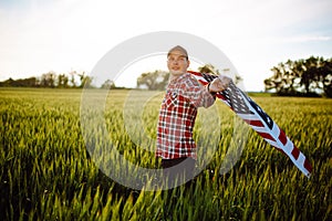 Young patriotic farmer stands among new harvest. Boy walking with the american flag on the green wheat field celebrating national