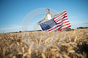 Young patriotic farmer stands among new harvest. Boy walking with the american flag on the wheat field celebrating