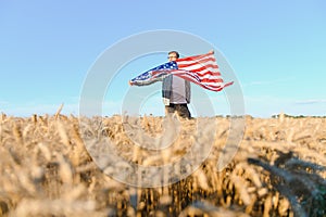 Young patriotic farmer stands among new harvest. Boy walking with the american flag on the wheat field celebrating