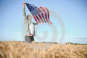 Young patriotic farmer stands among new harvest. Boy walking with the american flag on the wheat field celebrating