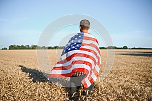 Young patriotic farmer stands among new harvest. Boy walking with the american flag on the wheat field celebrating