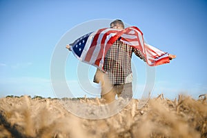 Young patriotic farmer stands among new harvest. Boy walking with the american flag on the wheat field celebrating