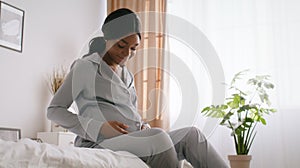 Young peaceful african american pregnant woman caressing her belly and smiling, sitting on bed in morning, empty space