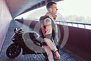 Young pensive biker is standing near his motobike in quiet tunnel