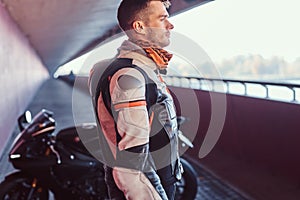 Young pensive biker is standing near his motobike in quiet tunnel