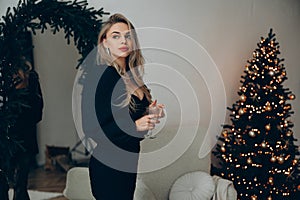 Young pensive blonde woman standing in decollete dress with wine glass in her hands near Christmas tree