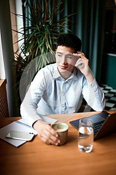 Young pensive business man wearing classic clothes working at modern city cafe with laptop and papers on desk. Freelance