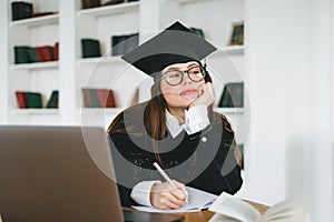 Young pensive caucasian woman college student in graduation outfit , sitting in front laptop with books and thinking about future