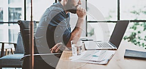 Young pensive coworker working at sunny work place loft while sitting at the wooden table.Man analyze document on laptop