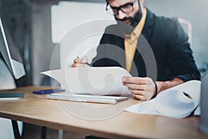 Young pensive coworker working at sunny work place loft while sitting at the wooden table.Man analyze paper document