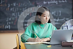 Young pensive female student in green pullover looking at laptop screen