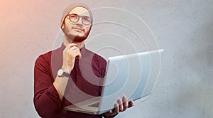 Young pensive guy holding laptop in hands over white background. Dressed in shirt and silver hat, wearing glasses.