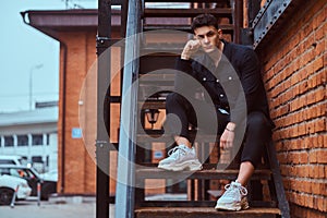 A young pensive guy sitting on stairs outside near a building with the industrial exterior.