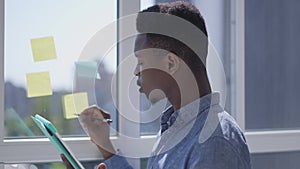Young pensive handsome African American startuper analyzing e-market on tablet writing ideas on sticky notes on window