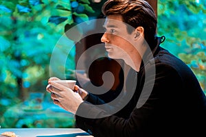 Young pensive handsome man drinking tea or coffee on rainy day and looking through the window.