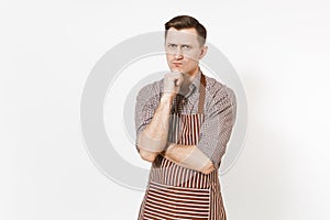 Young pensive man chef or waiter in striped brown apron, shirt holding hand to chin isolated on white background. Male