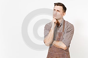 Young pensive man chef or waiter in striped brown apron, shirt holding hand to chin isolated on white background. Male