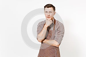 Young pensive man chef or waiter in striped brown apron, shirt holding hand to chin isolated on white background. Male