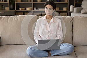 Young pensive serious woman sit on sofa working on laptop