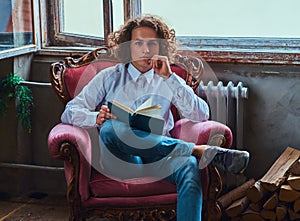 Young pensive student guy with curly hair holds a book and sitting on a chair.