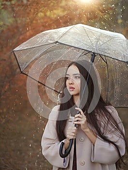 Young pensive woman in a coat under a transparent umbrella. The rain is falling and the sun is shining.