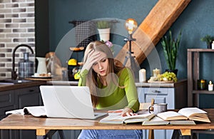 Young pensive woman freelancer sitting at table at home with laptop, making notes